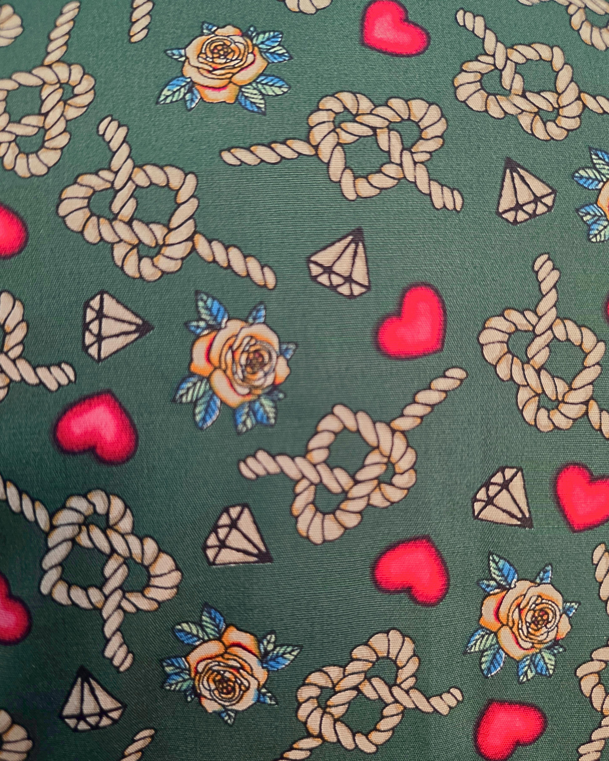 A close-up of a fabric pattern featuring beige rope knots, red hearts, tan roses with blue leaves, and small diamond motifs on a dark green background. Ruby De Vine ladies blouse handmade on the Gold Coast featuring a slim-fit stretch cotton design with short sleeves and a classic collar.